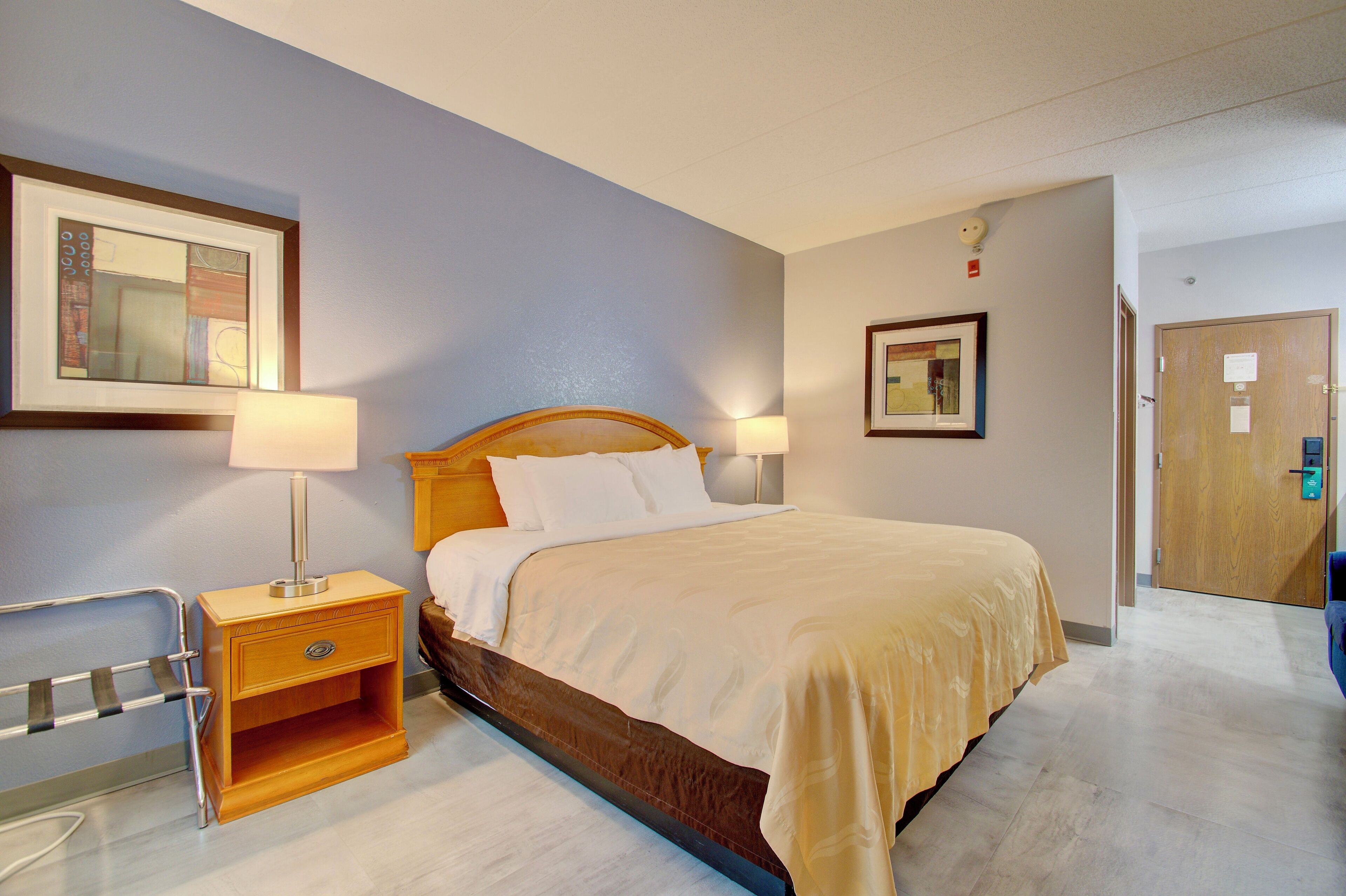 A hotel room with a bed, bedside lamps, a wooden nightstand, a framed picture on the wall, and a door with a lock.
