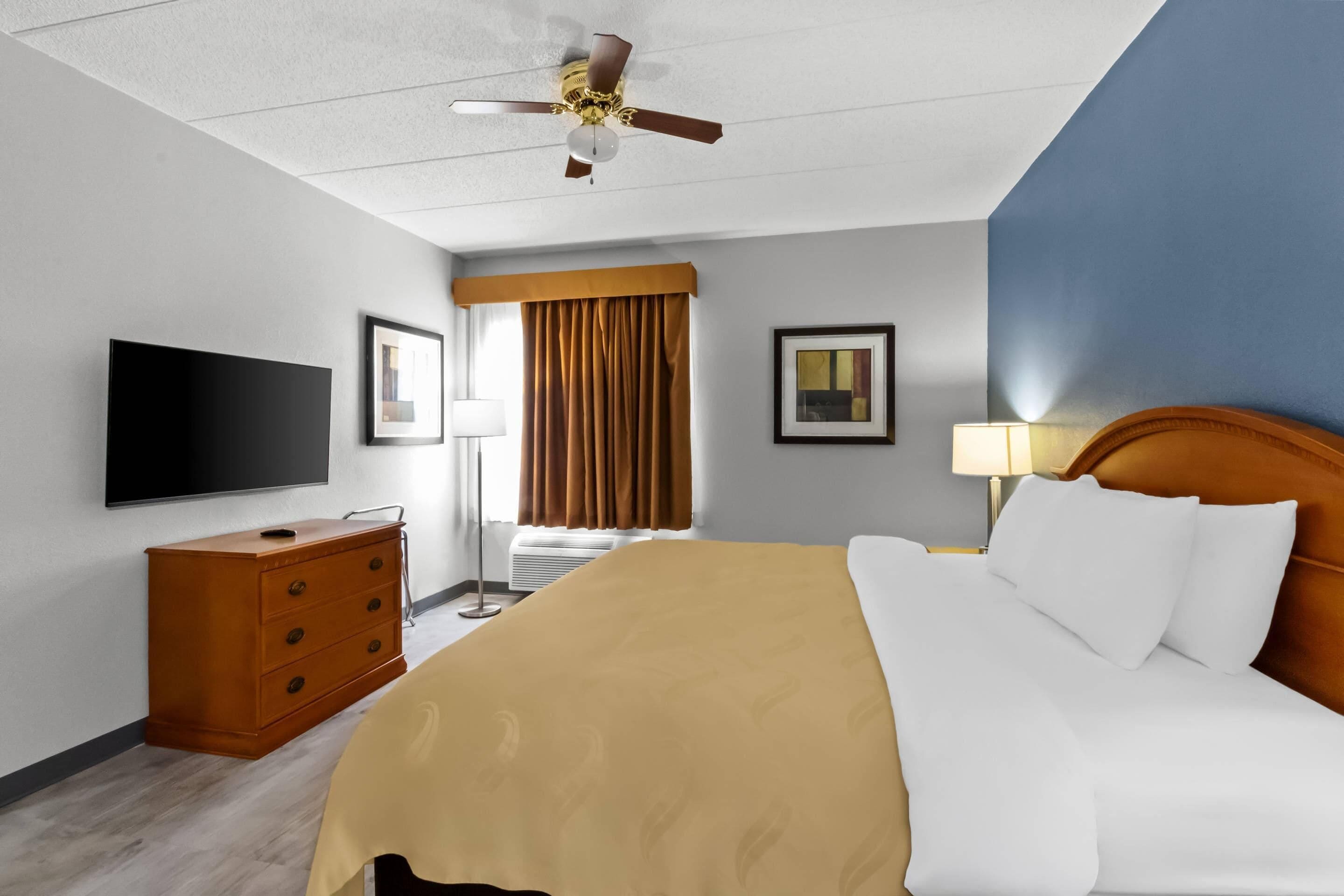 Habitación de hotel con cama, televisor, cómoda, ventilador de techo y ventana con cortinas.