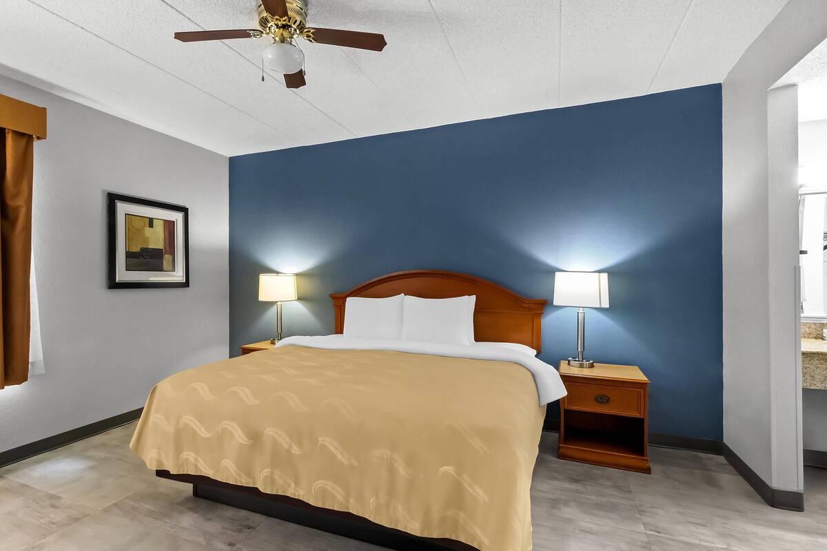 A hotel room with a bed, nightstands, a ceiling fan, a framed picture on the wall, and a window with curtains.