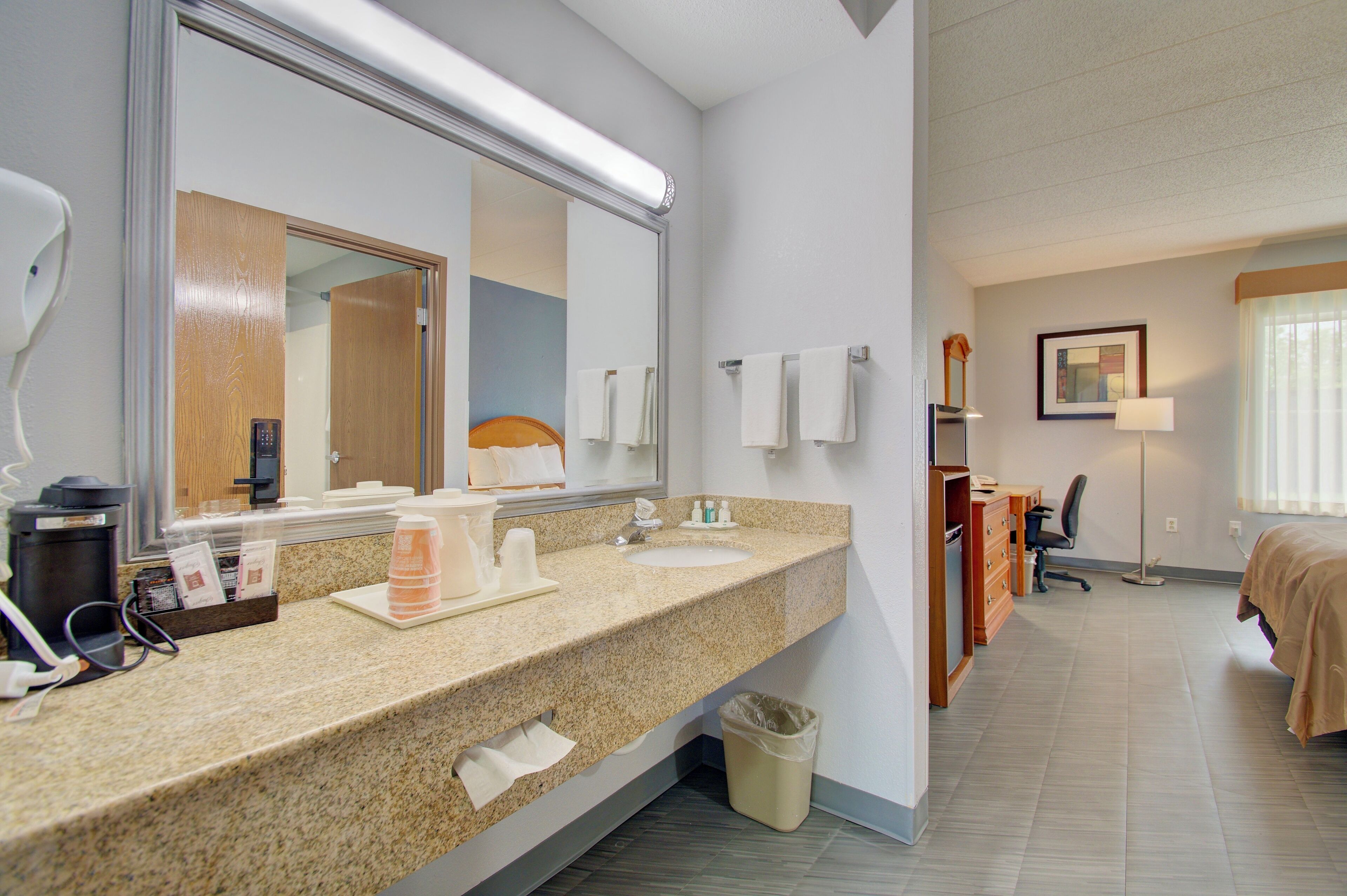 A hotel bathroom with a granite countertop, a sink, a mirror, a trash bin, and a wooden vanity.