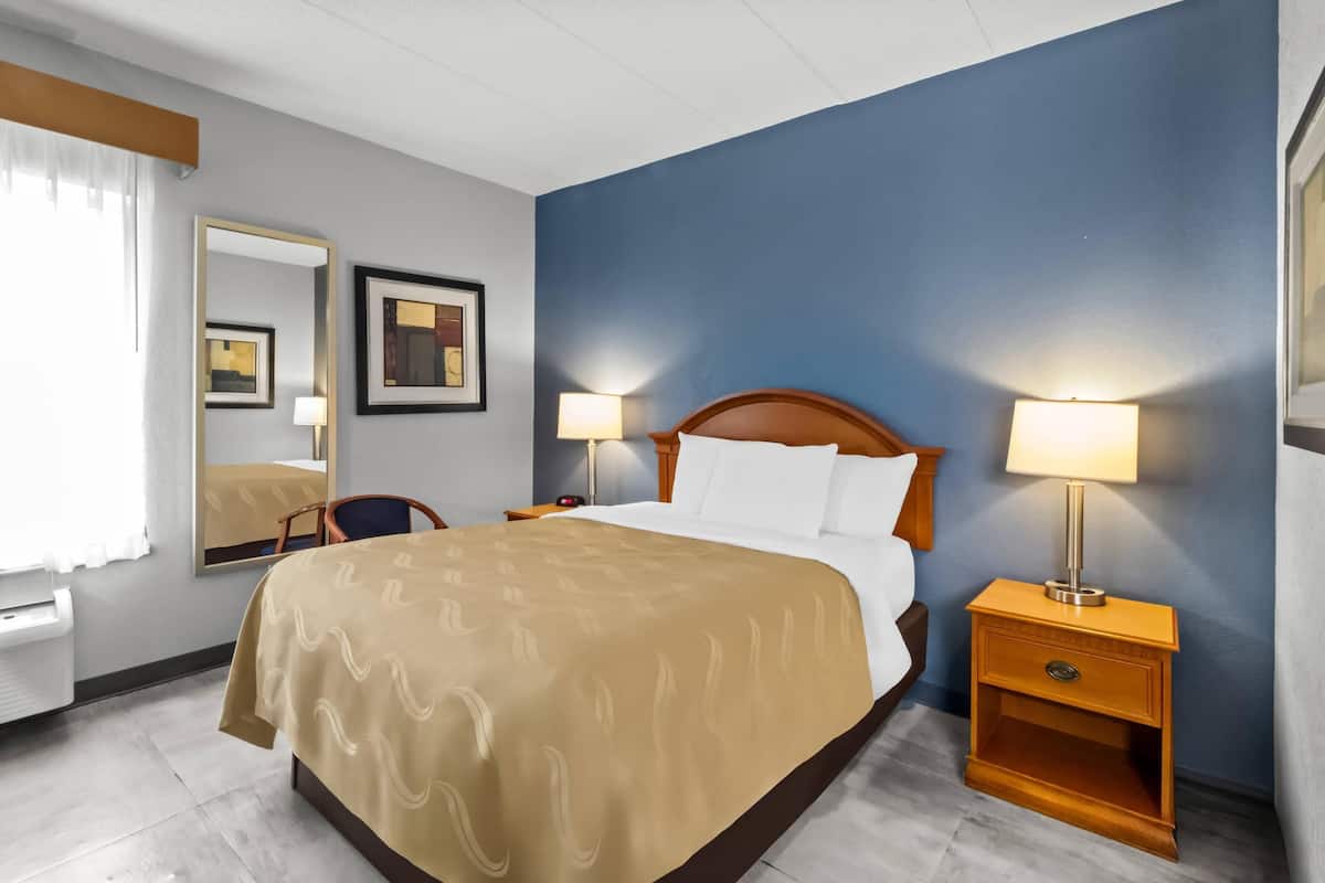 A hotel room with a bed, bedside lamps, a nightstand, a chair, a mirror, and framed pictures on the wall.
