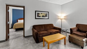 A hotel room with a brown sofa, a wooden coffee table, a small round table, a lamp, and a framed picture on the wall. - Quality Inn & Suites CVG Airport (Hebron)