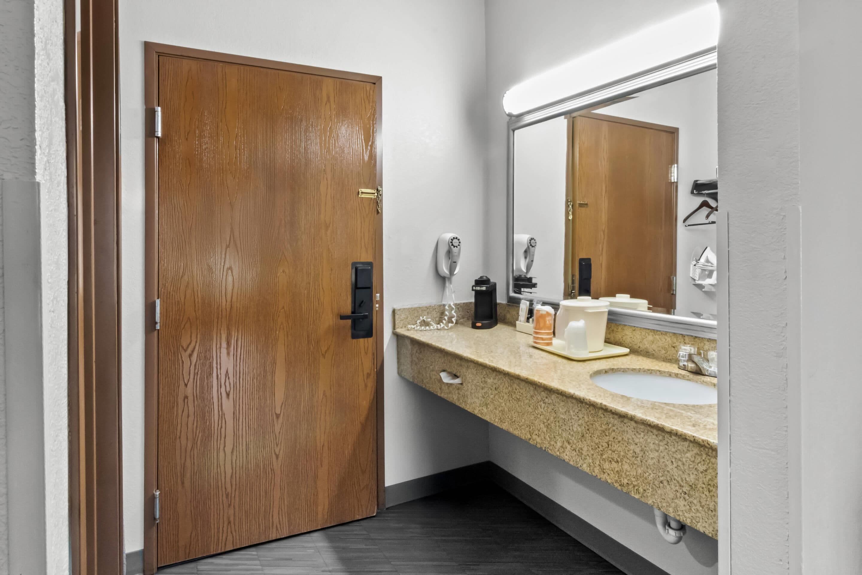 Un baño con puerta de madera, lavamanos, espejo, secador de pelo y cafetera.