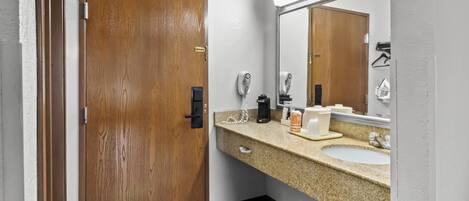 A bathroom with a wooden door, a sink, a mirror, a hairdryer, and a coffee maker.
