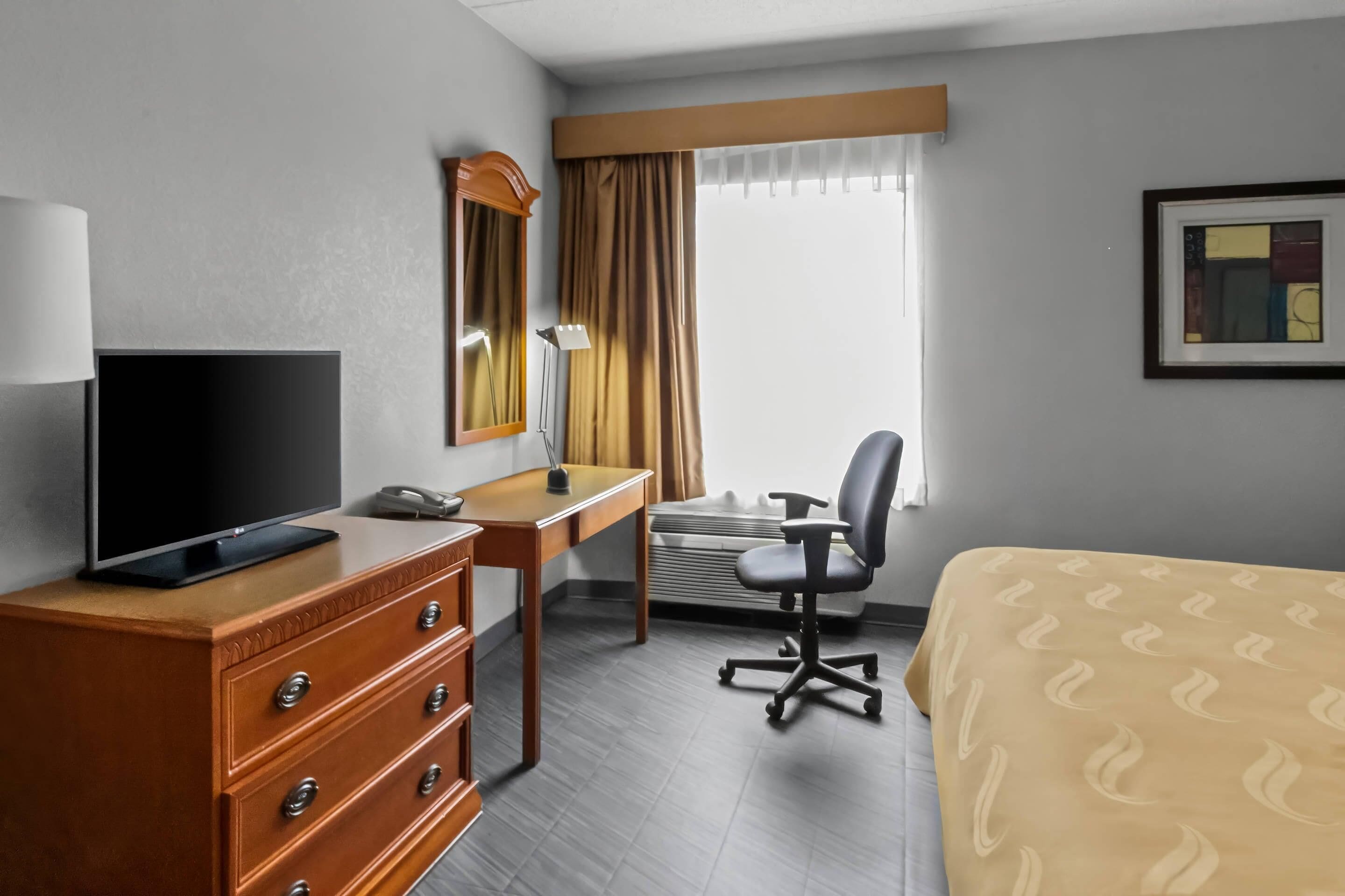 A hotel room with a TV on a dresser, a desk with a chair, a bed, a window with curtains, and a framed picture on the wall.