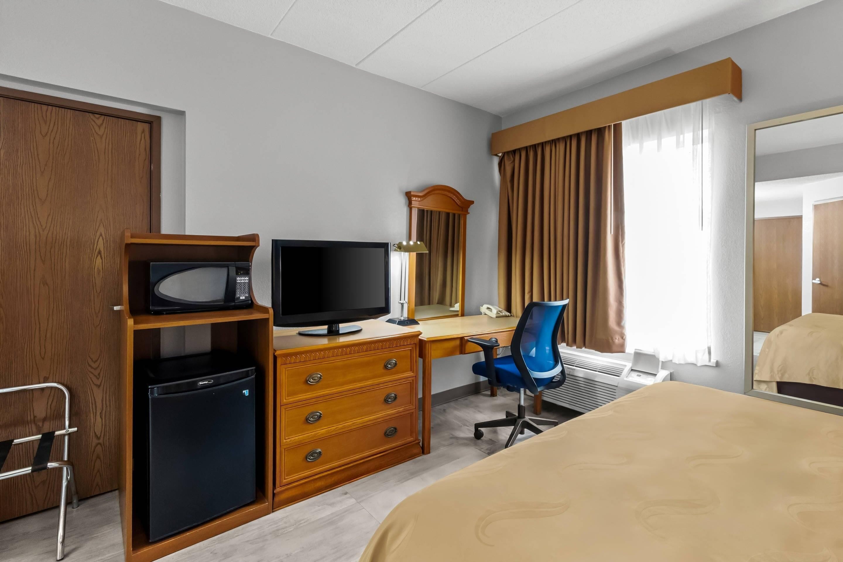 A hotel room with a bed, a desk with a computer, a chair, a television, a microwave, a refrigerator, a mirror, and a window with curtains.