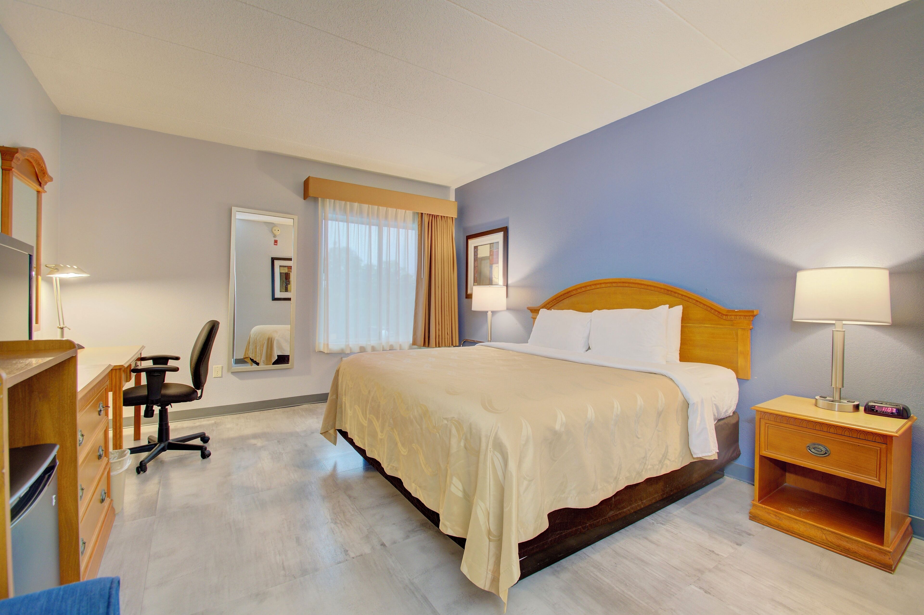 A hotel room with a bed, a desk, a chair, a nightstand, and a mirror.