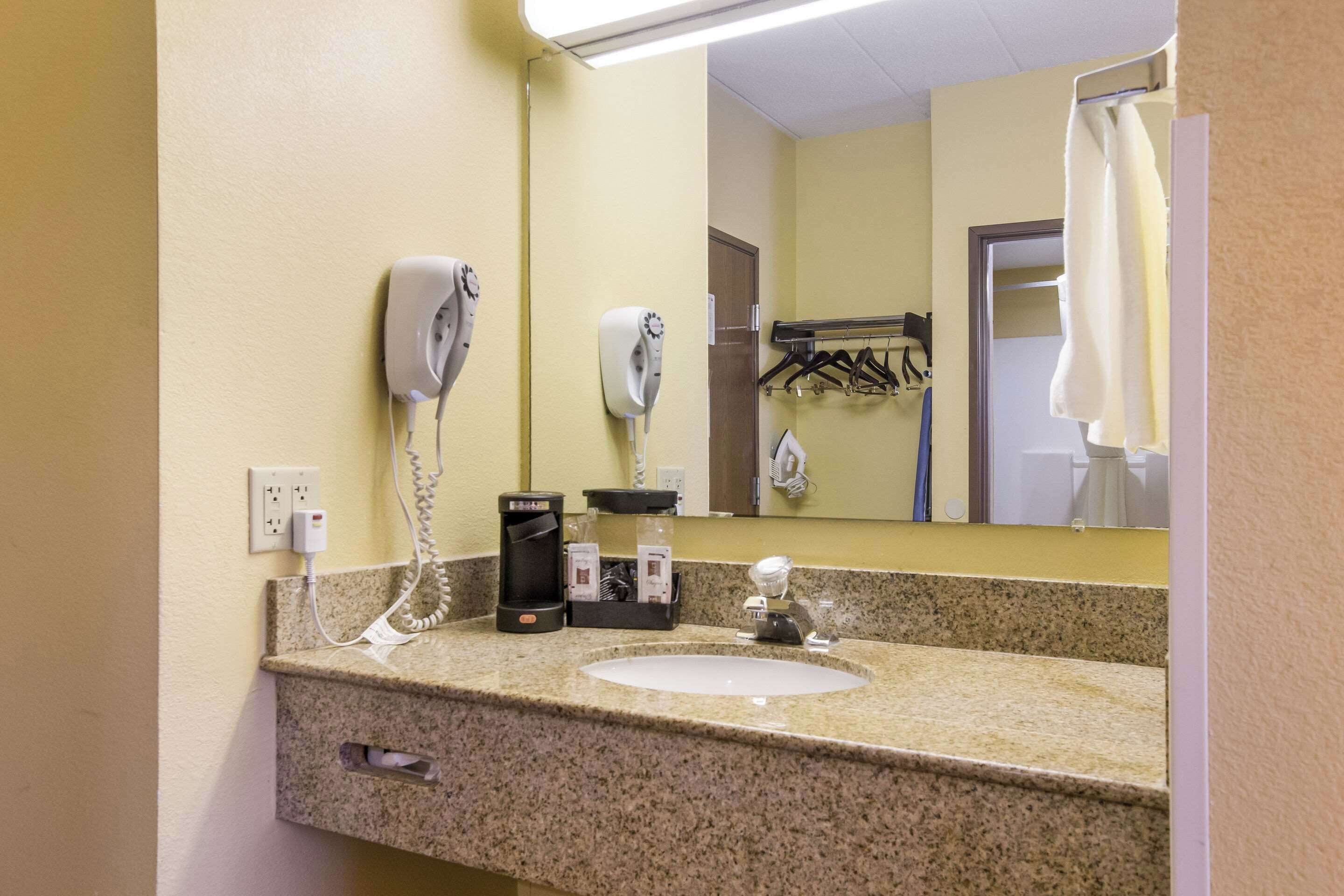 Un baño de hotel con encimera de granito, un lavamanos, un espejo, un secador de pelo, un perchero para toallas y una cafetera.