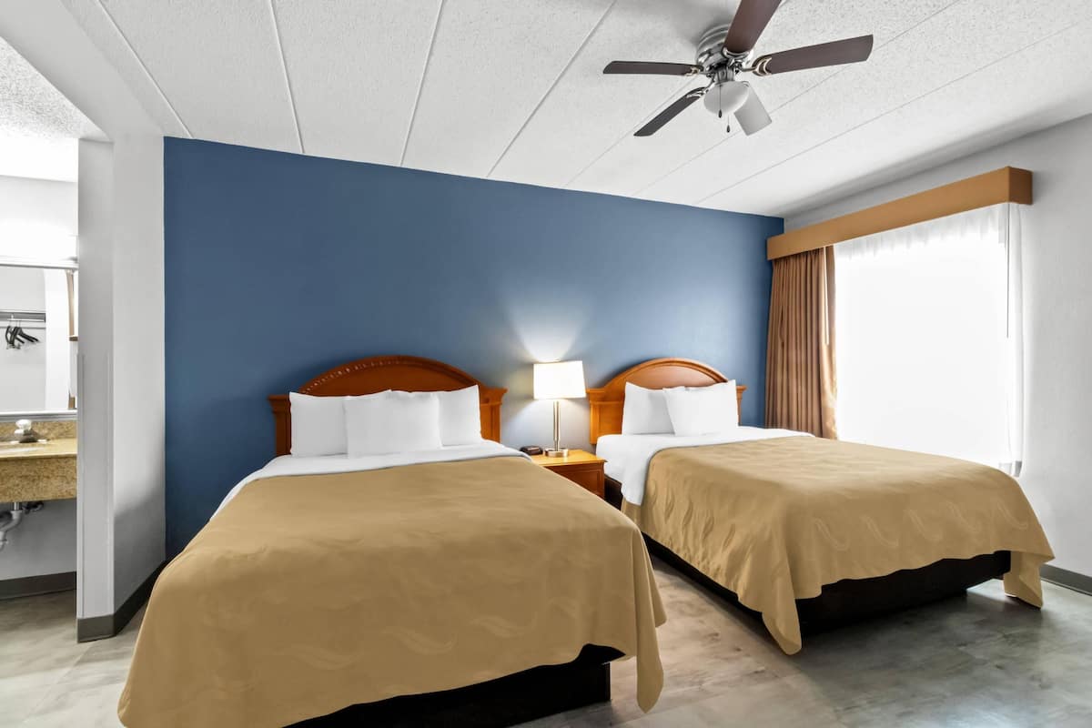 A hotel room with two beds, a ceiling fan, a nightstand with a lamp, and a large window with curtains.