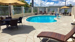 Zona de piscina con sillones y sombrillas, rodeada por una valla y edificios al fondo.