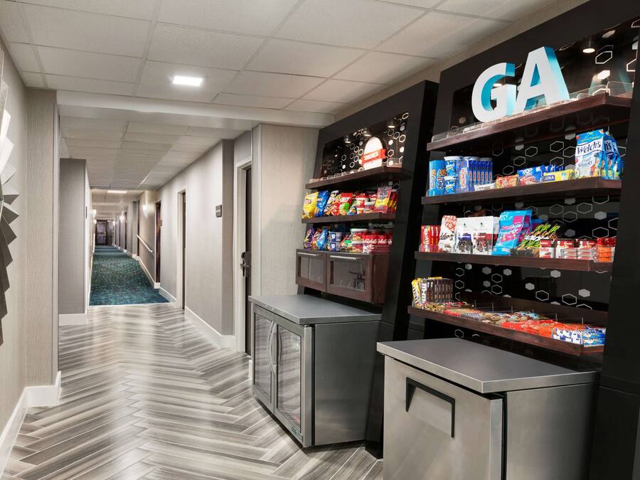 A hotel corridor with a herringbone-patterned floor, a vending machine, and shelves stocked with snacks and beverages.