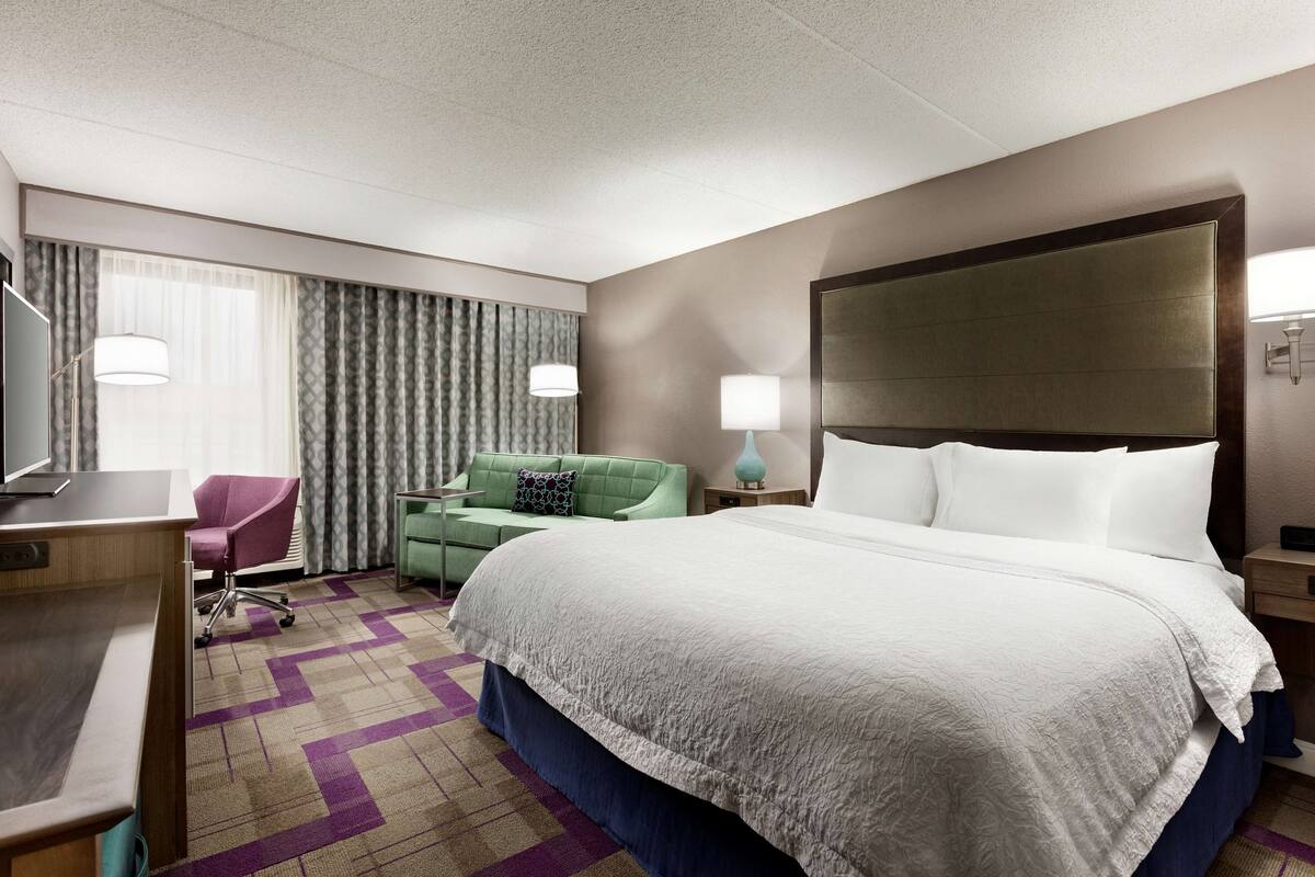 A hotel room with a large bed, a desk, a green armchair, and a TV.