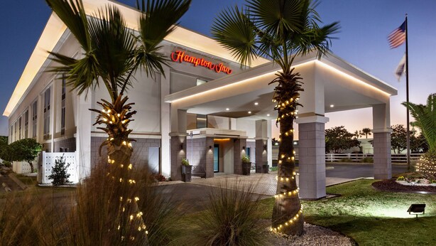 Un hotel Hampton Inn con diseño moderno, con palmeras y una entrada bien iluminada.