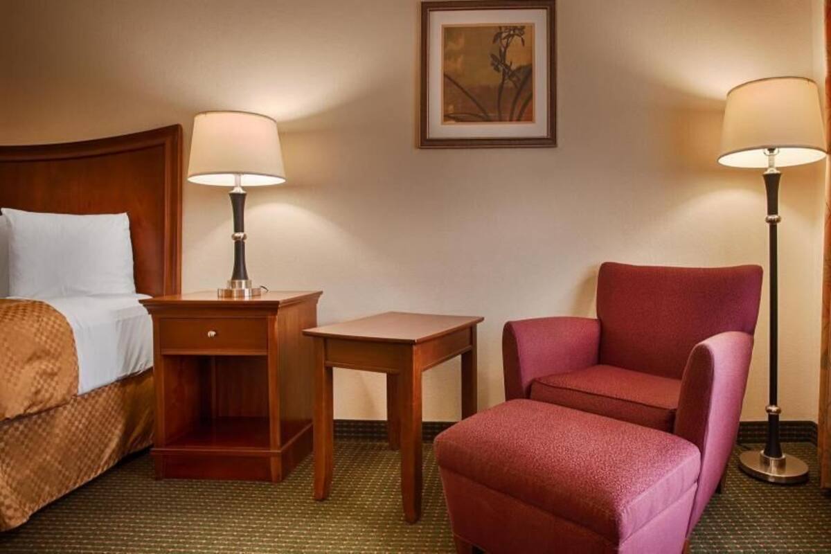 Hotellihuone, jossa on sänky, työpöytä, tuoli, pöytä, kaksi lamppua, taulu seinällä ja ikkuna, jossa on verhot.