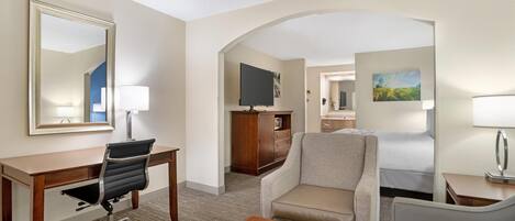 Habitación de hotel con escritorio, silla de oficina, dos sillones, cama, televisor, cómoda y un cuadro en la pared.