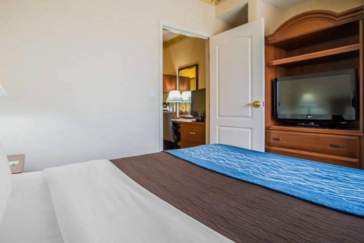 Quarto de hotel com uma cama, televisão, escrivaninha com luminária e porta que dá para outro cômodo.