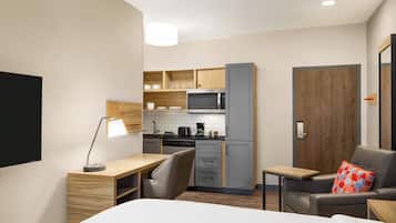 A compact hotel room with a kitchenette, a bed, a desk, and a seating area.