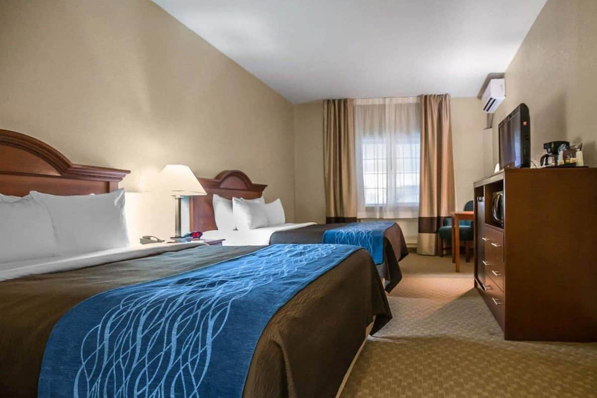 Quarto de hotel com duas camas, uma escrivaninha, uma cadeira, uma televisão e um guarda-roupa.