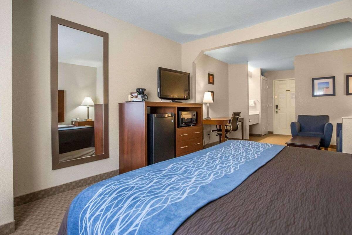 A hotel room with a large bed, a desk with a chair, a television, a refrigerator, and a mirror.