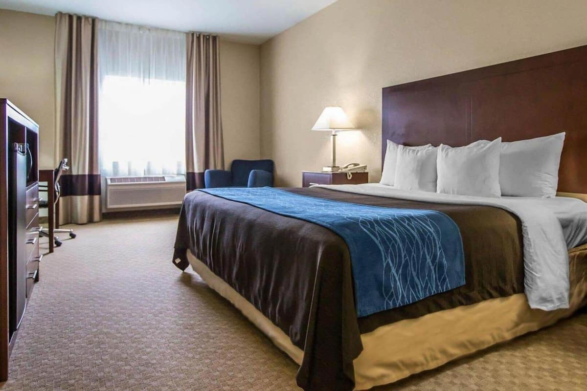 Quarto de hotel com uma cama grande, uma escrivaninha, uma cadeira, um abajur e vista para o exterior pela janela.