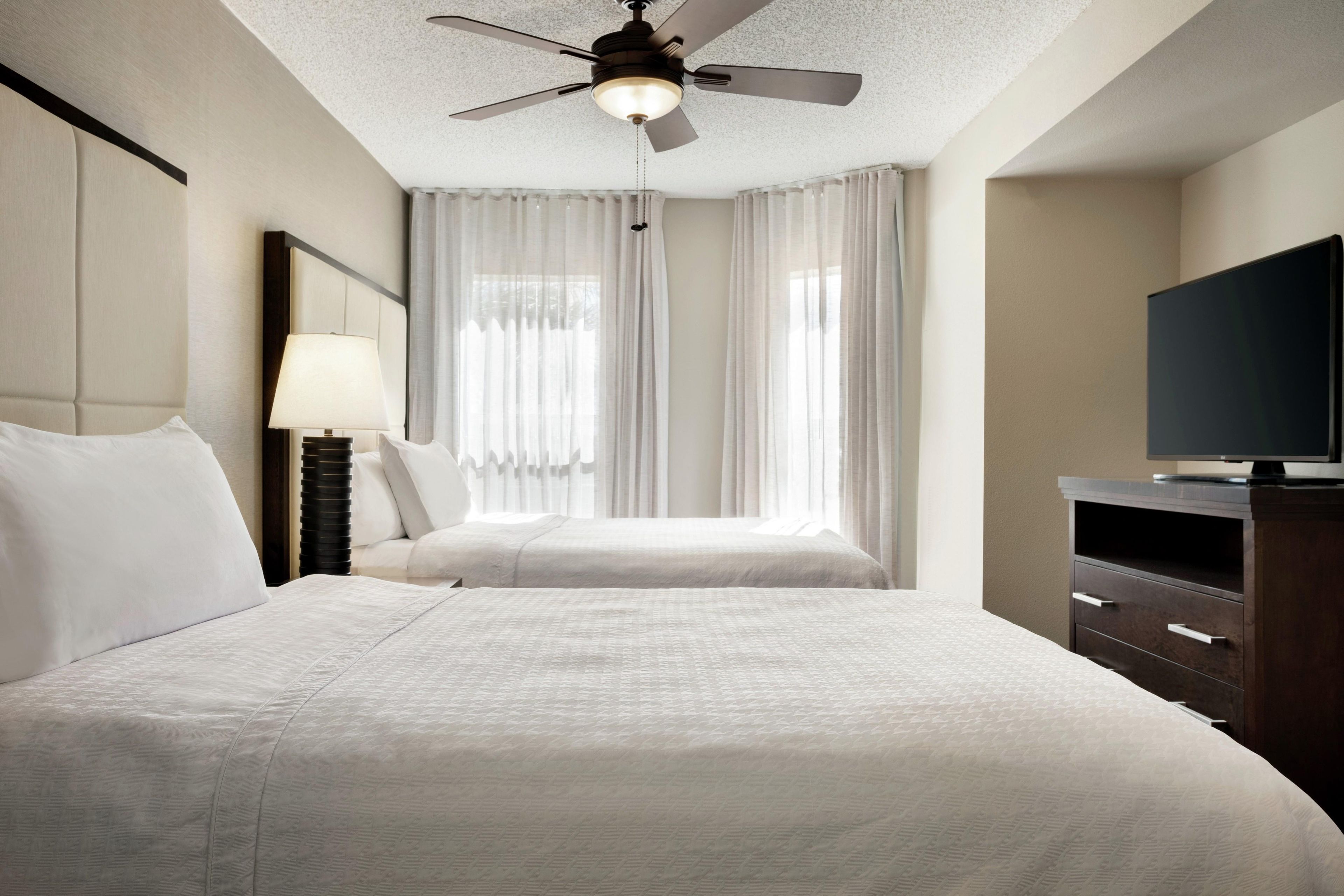 A hotel room with two beds, a ceiling fan, a TV, and a nightstand with a lamp.