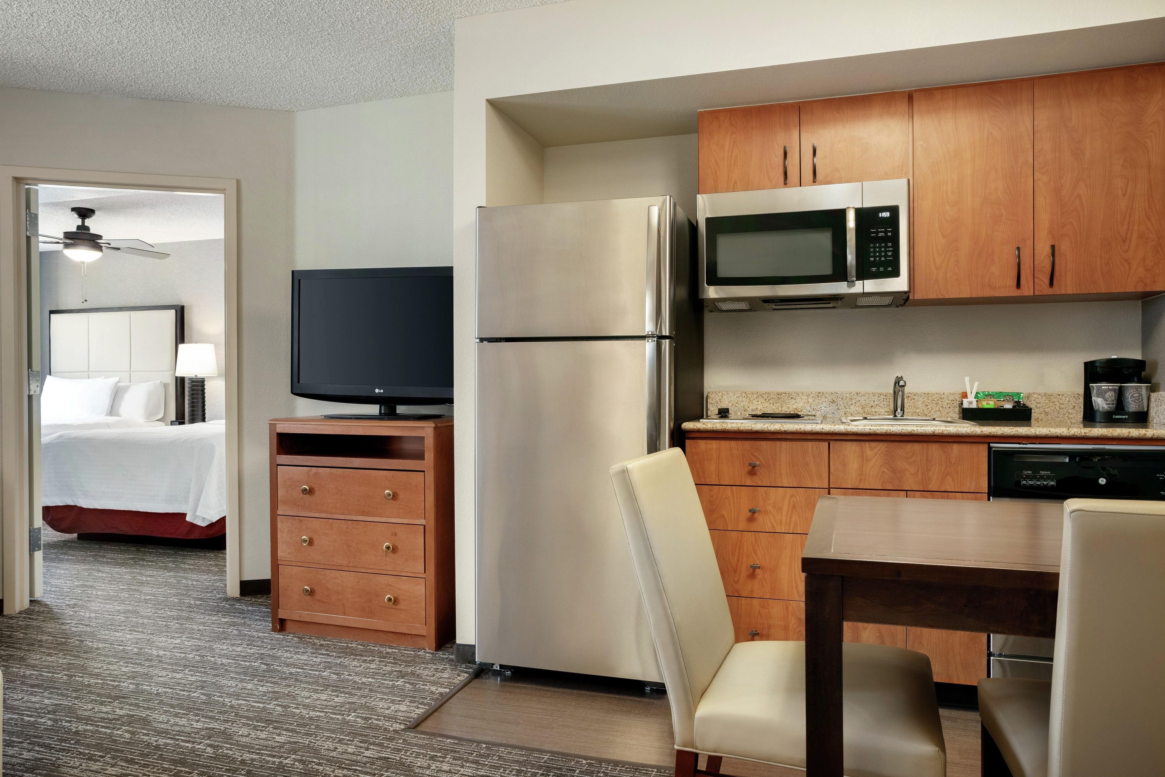 Habitación de hotel con cama, televisión, refrigerador, microondas, armario de madera y zona de comedor con mesa y sillas.