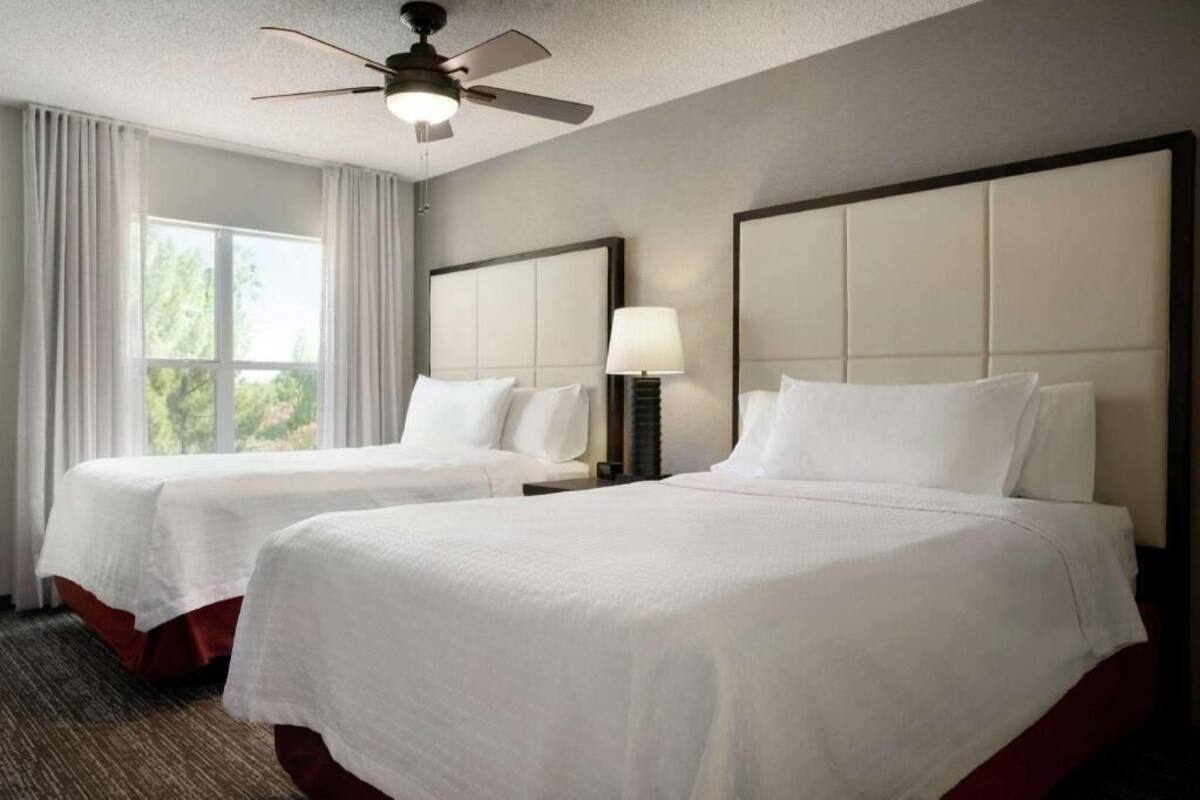 A hotel room with two beds, a ceiling fan, a window with curtains, and a nightstand with a lamp.