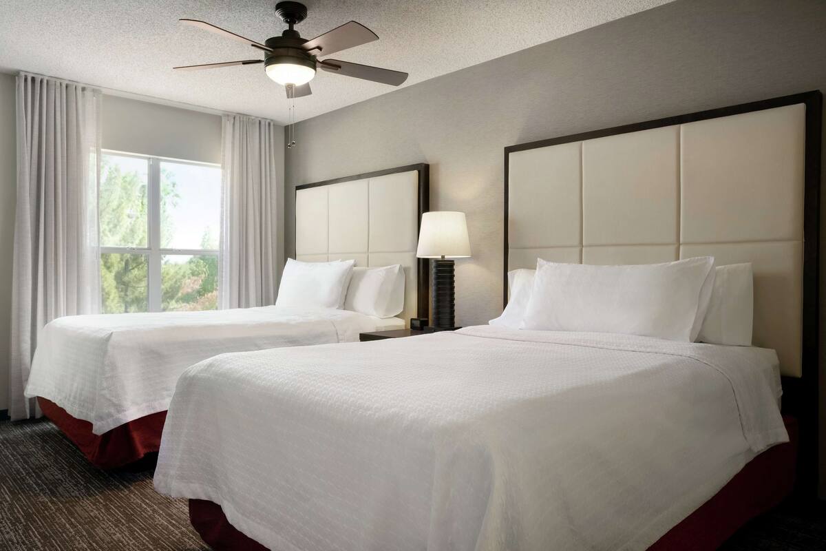 A hotel room with two beds, a ceiling fan, a window with curtains, and a nightstand with a lamp.