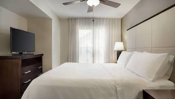 Ein Hotelzimmer mit Bett, Fernseher auf einem Schminktisch, Deckenventilator und einem Fenster mit Vorhängen.