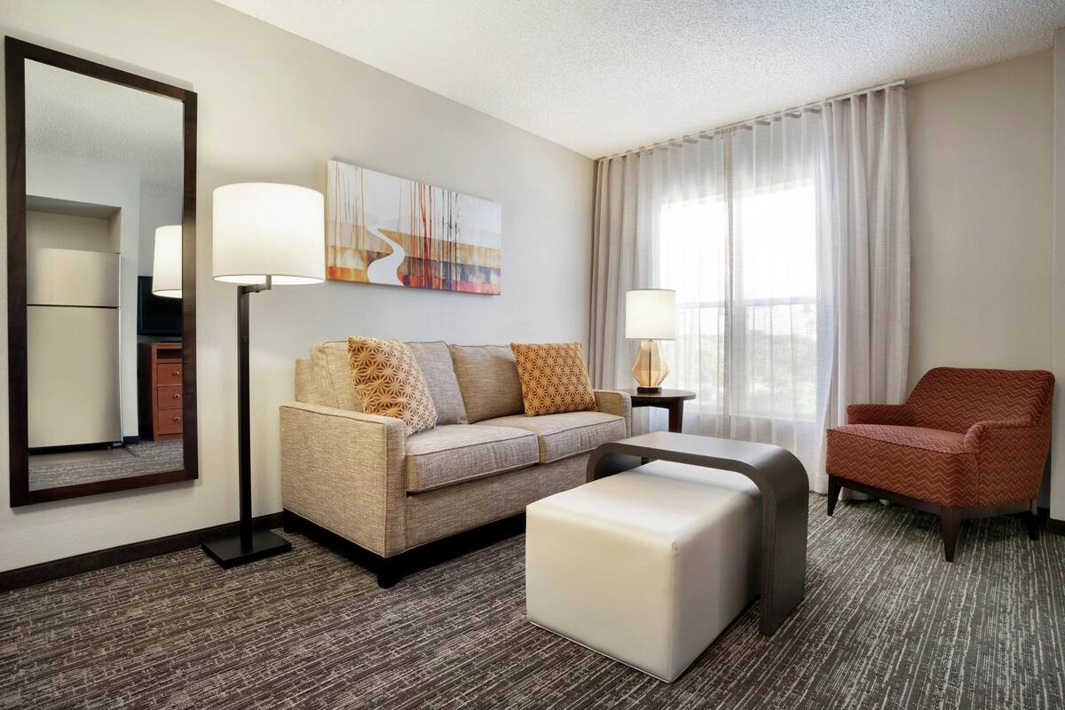 A hotel room with a sofa, ottoman, chair, lamp, and artwork on the wall.