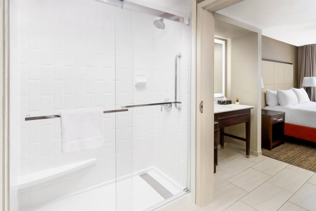 A hotel bathroom with a glass-enclosed shower, a sink, and a bed with a red accent.