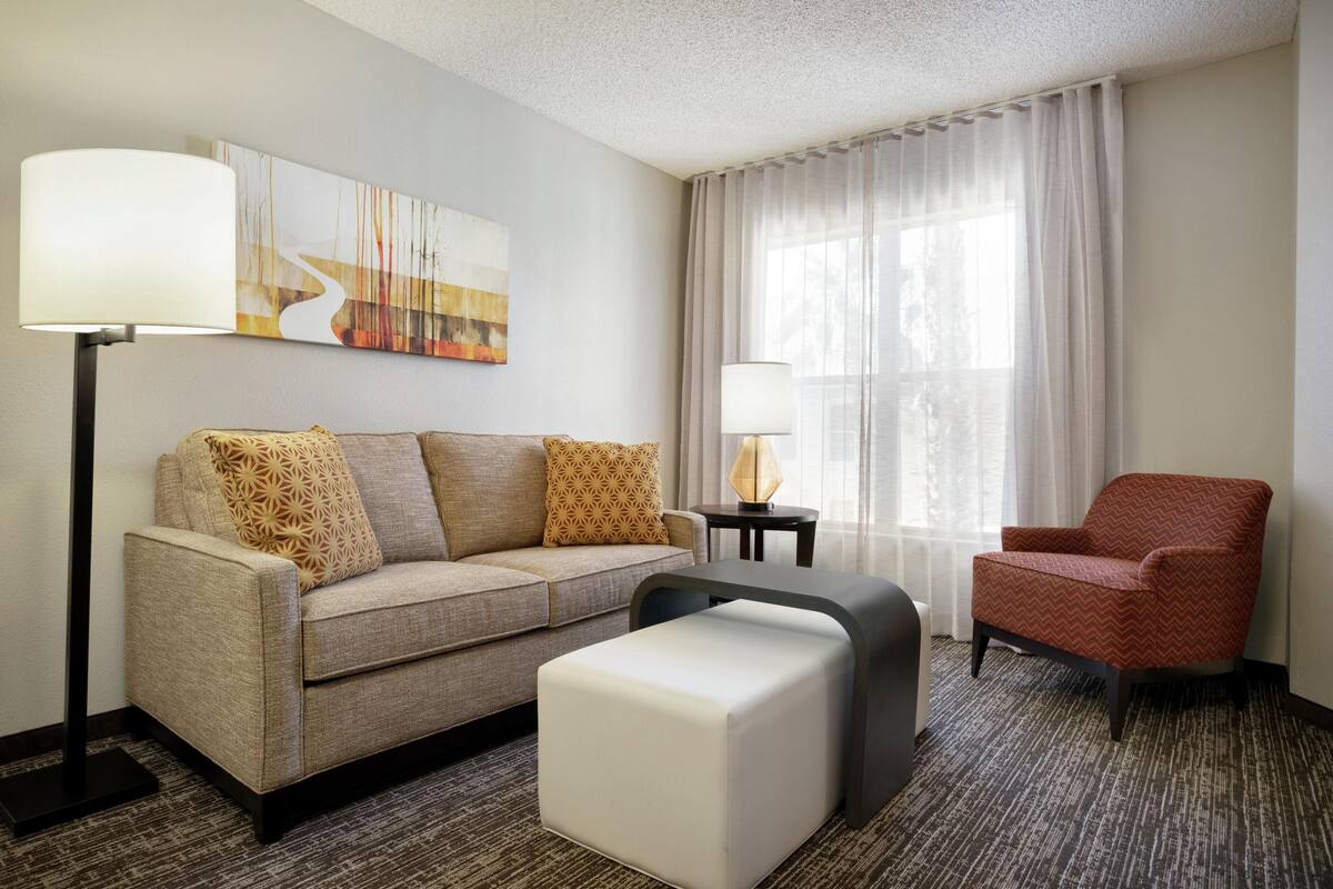 A hotel room with a sofa, ottoman, armchair, floor lamp, table lamp, and a painting on the wall.