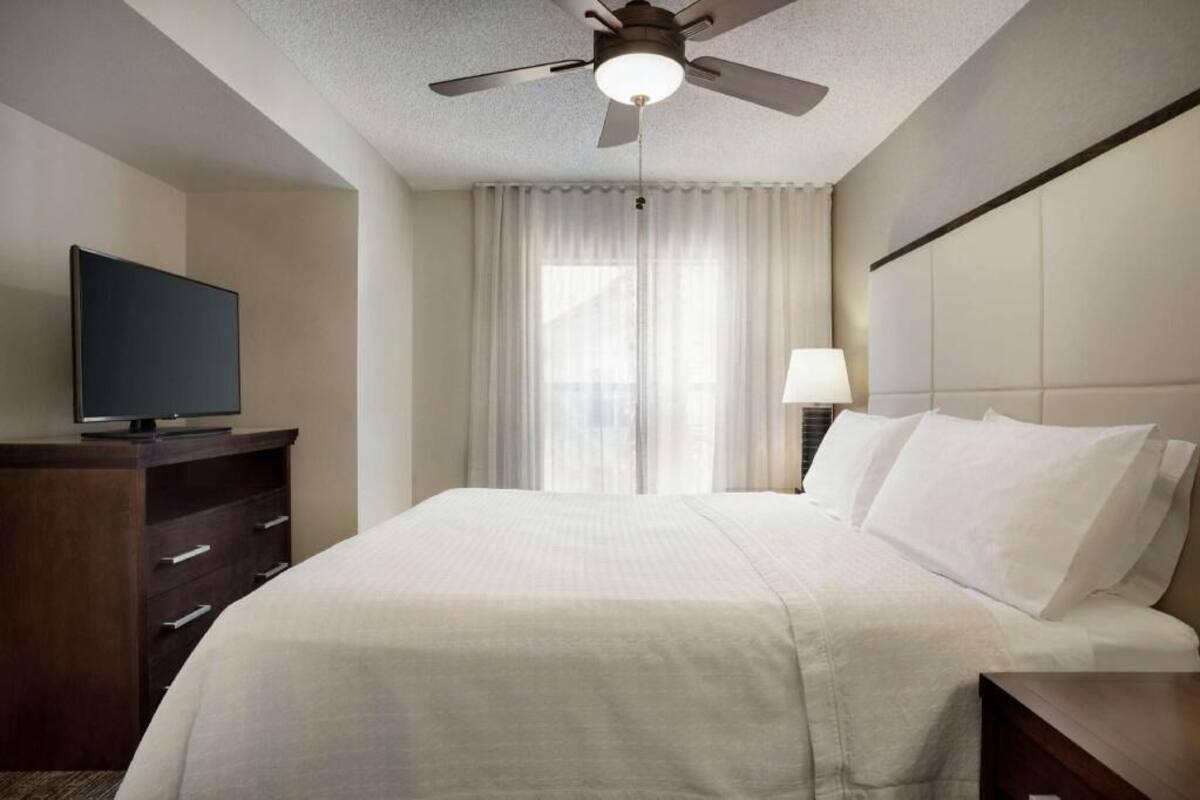 A hotel room with a bed, a TV on a dresser, a ceiling fan, a window with curtains, and a bedside table with a lamp.
