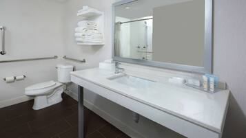 A modern bathroom with a white sink, mirror, and toilet. There are handrails on the walls and a shelf with towels.