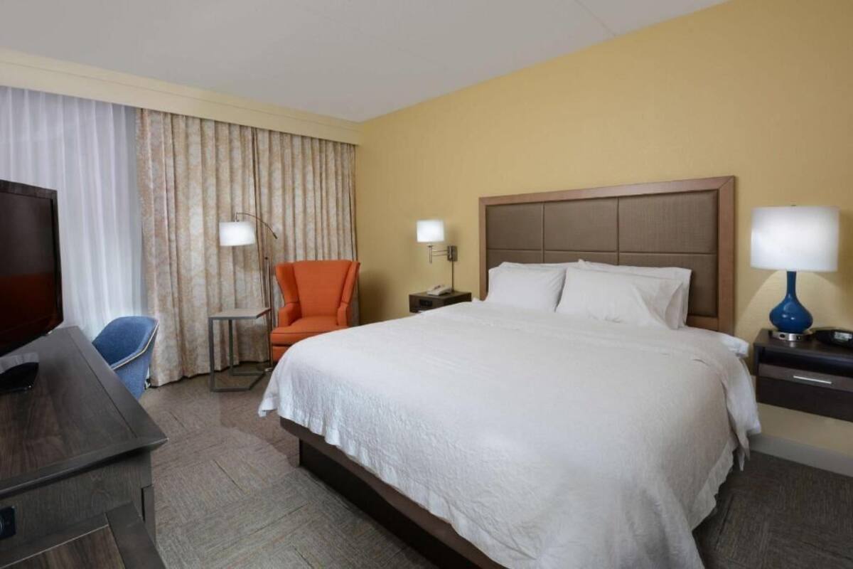 A hotel room with a bed, a desk, an orange chair, two bedside lamps, and a television.