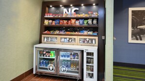 A snack display with various chips and snacks, a refrigerated beverage cooler, and a sign above the snacks. - Hampton Inn High Point (High Point)