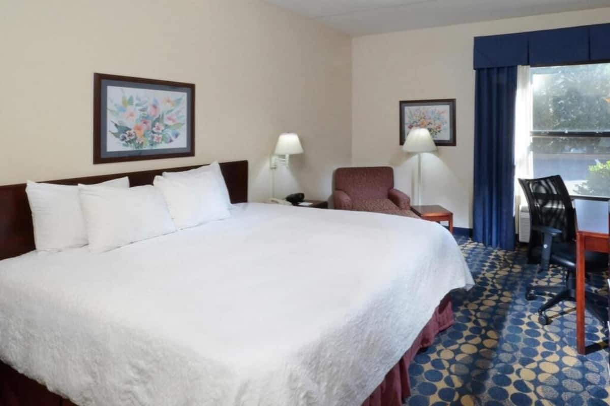 A hotel room with a large bed, two bedside lamps, a desk with a chair, and two framed floral artworks on the walls.