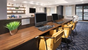 A modern hotel reception area with a wooden reception desk, two monitors, a keyboard, and a potted plant. There are chairs and a sofa in the background. - Courtyard by Marriott Burlington (Burlington)