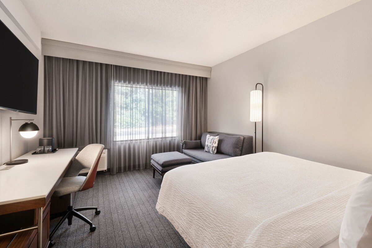 A hotel room with a bed, a desk with a chair, a sofa, a TV, and a window with curtains.