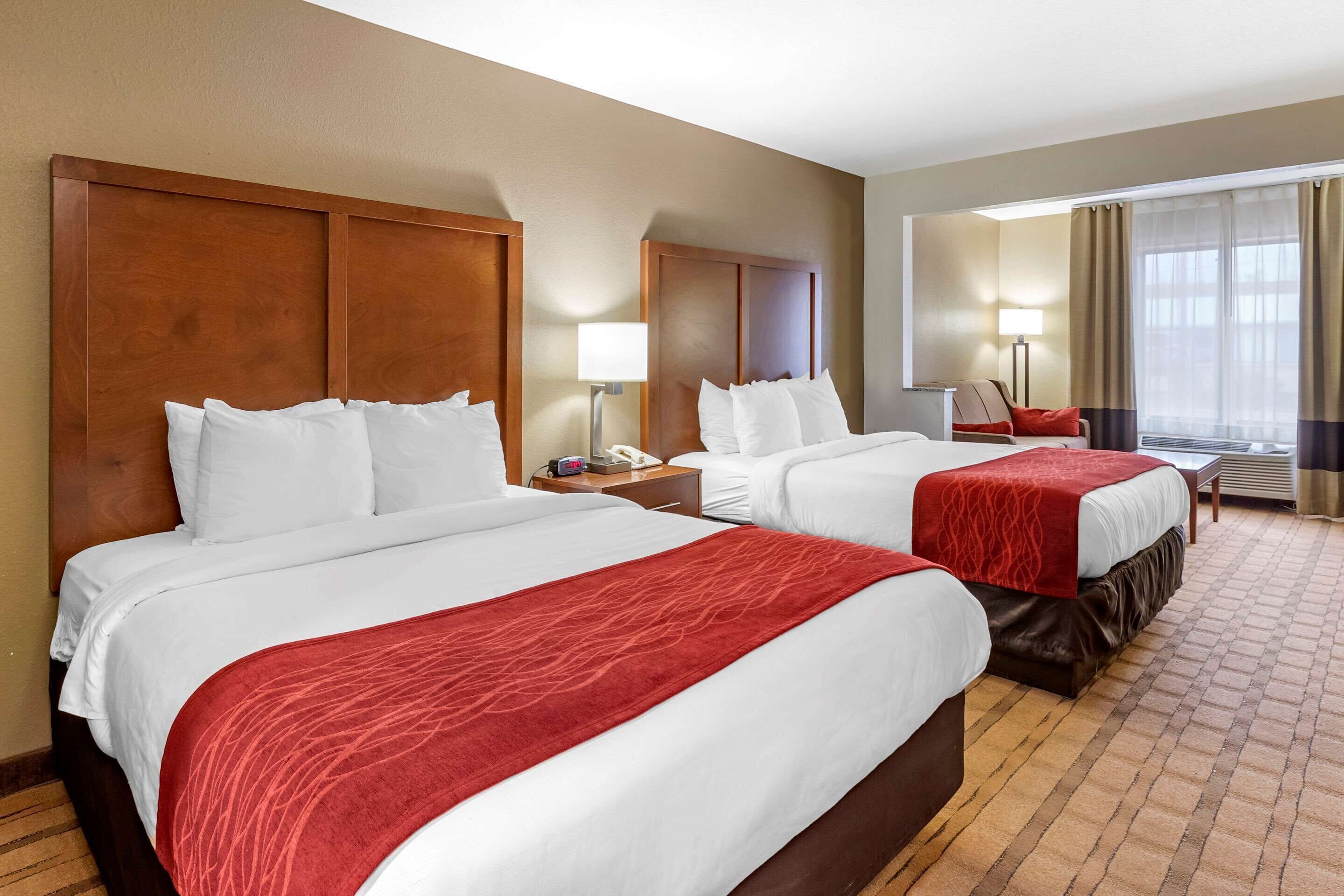Habitación de hotel con dos camas, cada una con sábanas blancas y mantas rojas, una mesita de noche con lámpara, un escritorio con silla y un ventanal con cortinas.