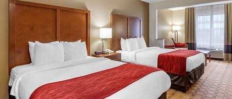 Habitación de hotel con dos camas, cada una con sábanas blancas y mantas rojas, una mesita de noche con lámpara, un escritorio con silla y un ventanal con cortinas.