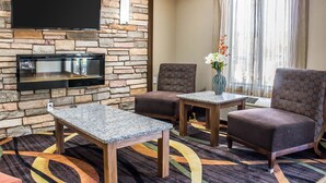 A hotel lobby with a stone wall, a flat-screen TV, two armchairs, a small table, and a colorful carpet. - Comfort Inn & Suites Cedar Rapids North - Collins Road (Cedar Rapids)