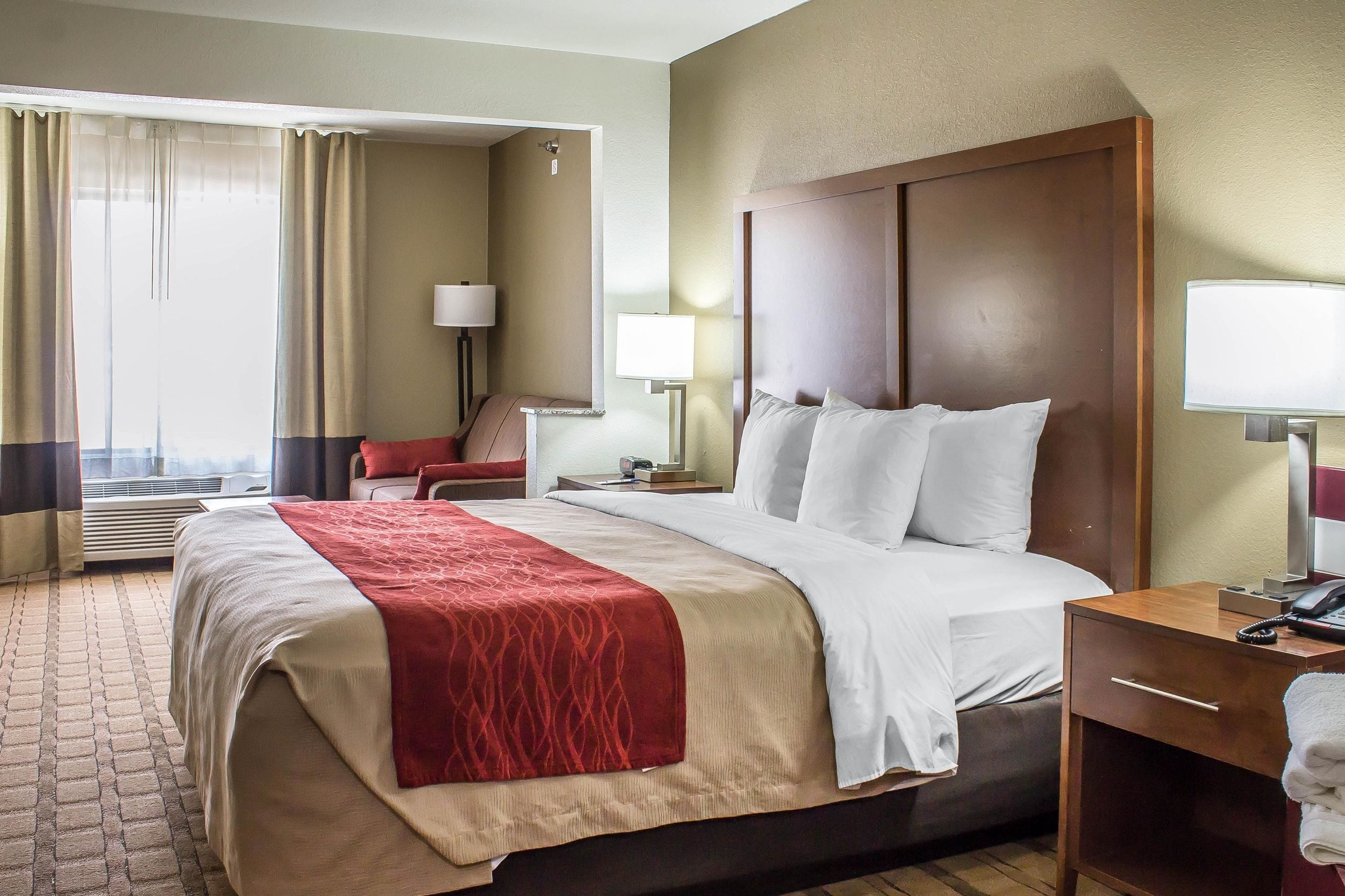 A hotel room with a large bed, a wooden headboard, a nightstand with a lamp, a chair, and a view of the city through the window.