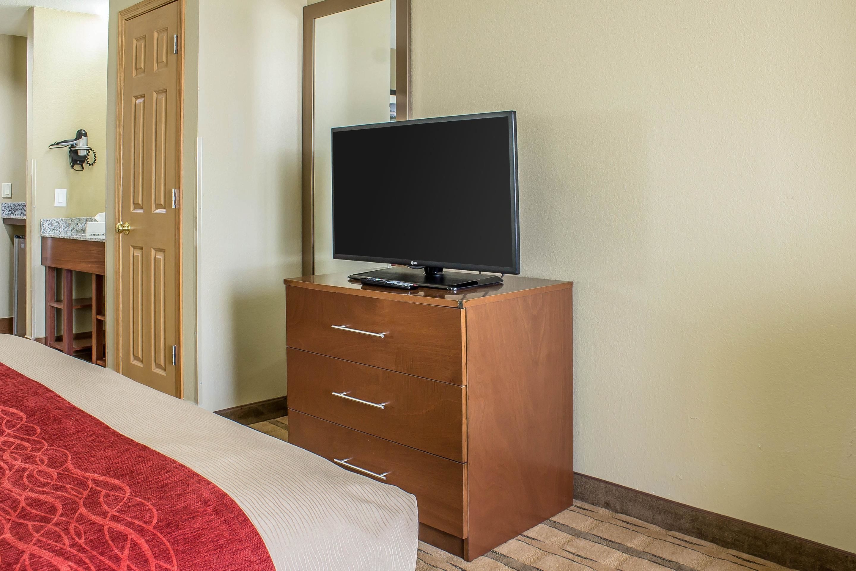 A hotel room with a wooden dresser, a flat-screen TV, a bed with a red and white bedspread, and a bathroom visible in the background.
