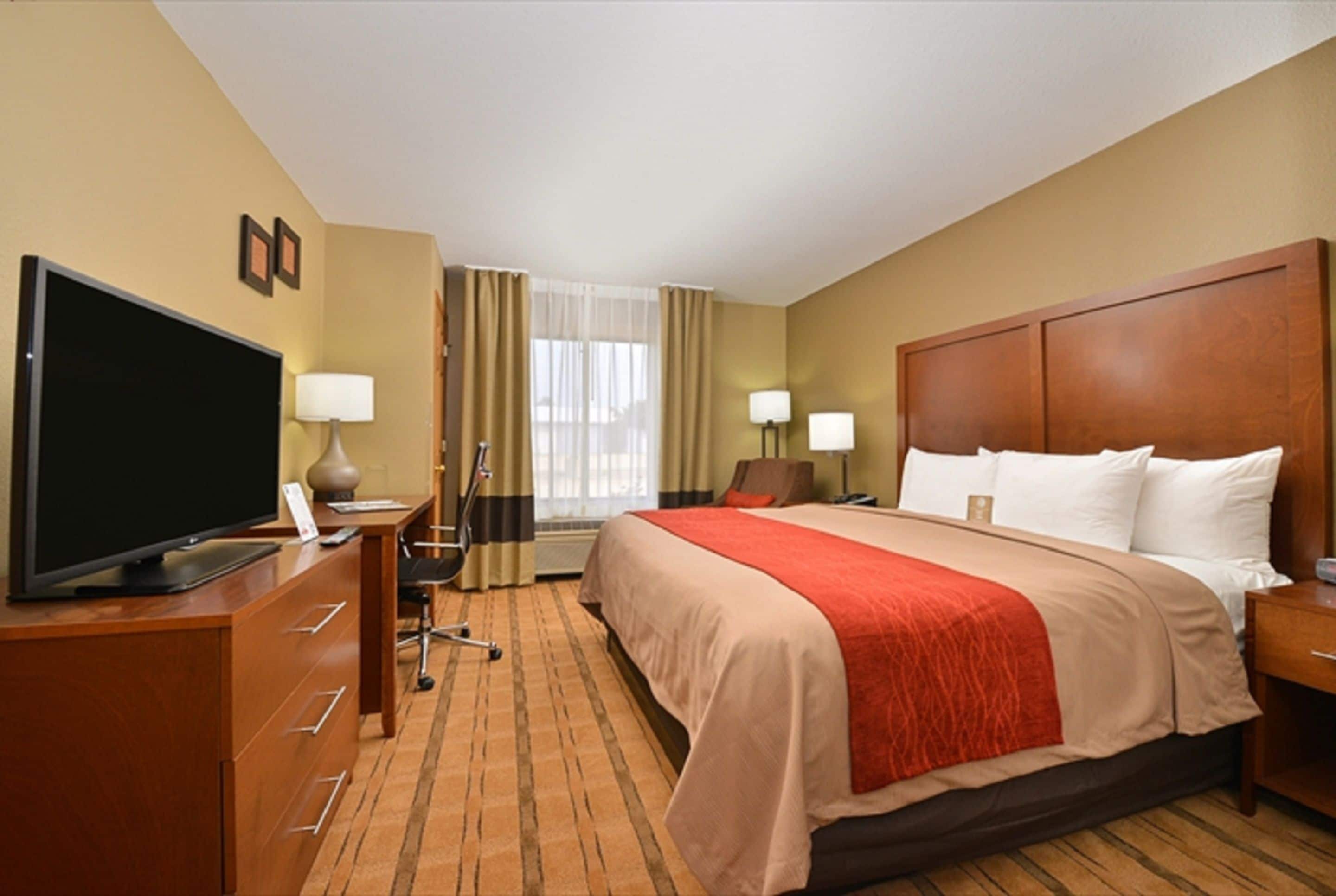 A hotel room with a large bed, a desk with a flat-screen TV, two bedside lamps, and a window with curtains.