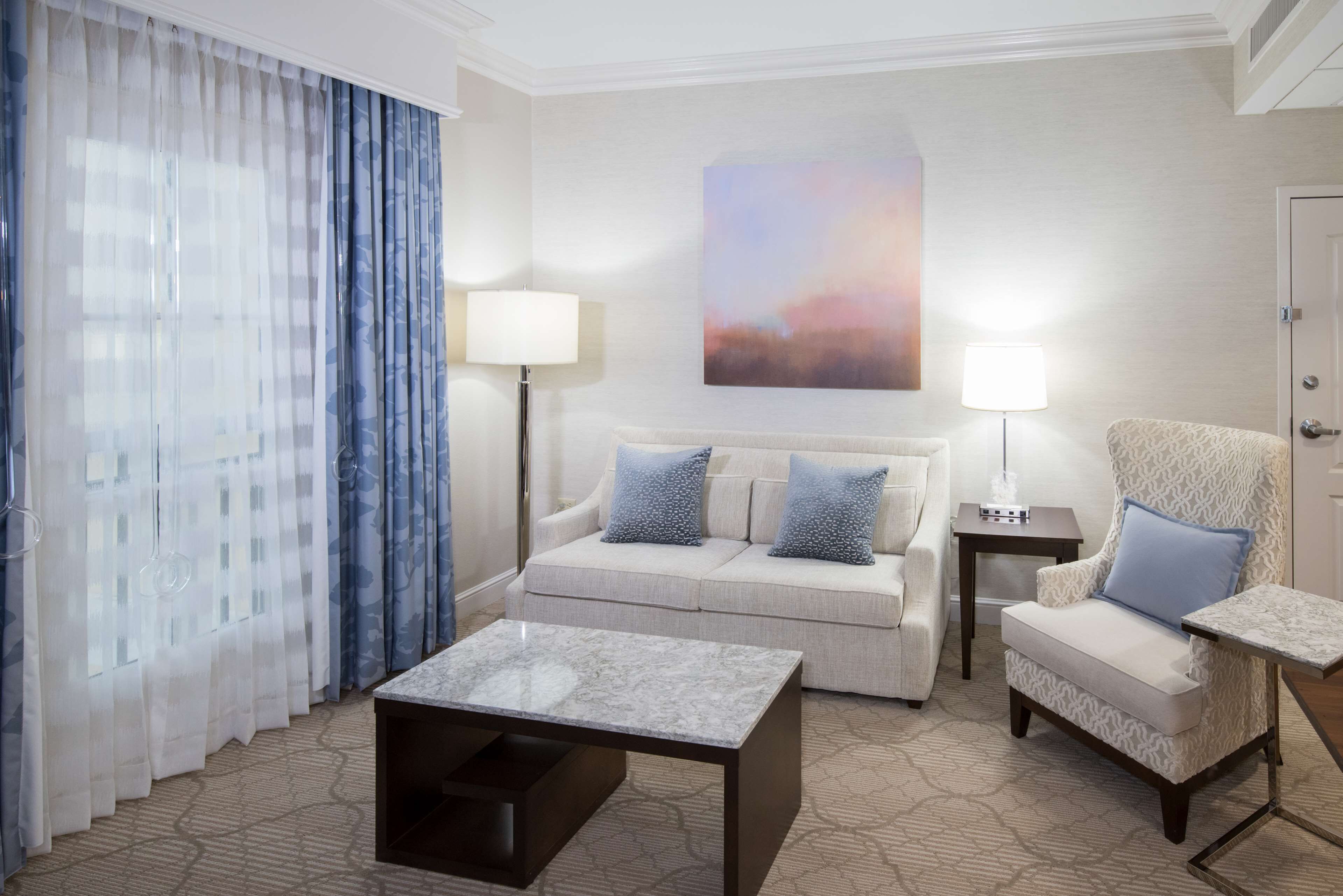 A hotel room with a sofa, armchair, coffee table, and a painting on the wall.