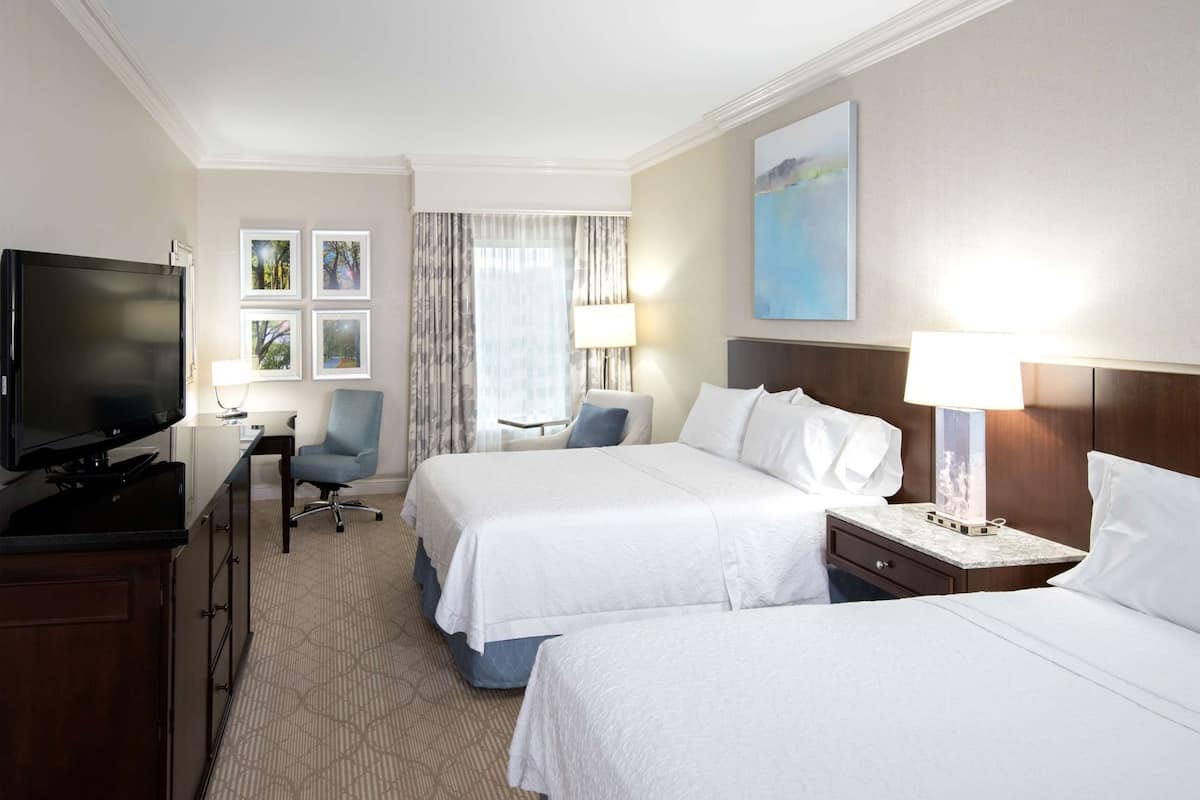 A hotel room with two beds, a desk with a television, a chair, a lamp, and artwork on the walls.