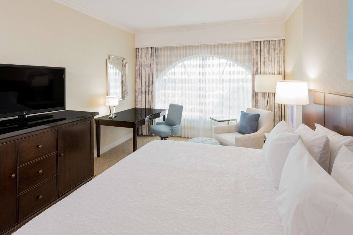 A hotel room with a large bed, a desk with a TV, a chair, a small table, and a lamp.