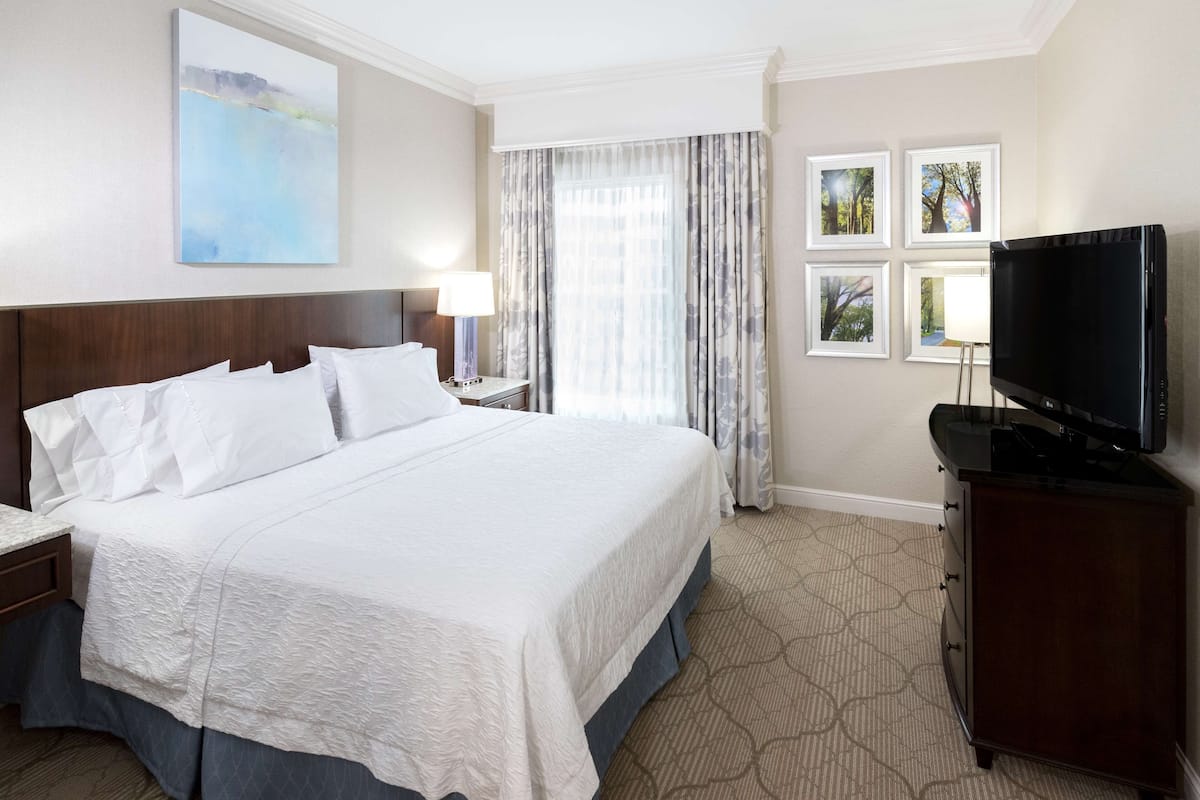 A hotel room with a large bed, a flat-screen TV, a nightstand, a lamp, and a window with curtains.