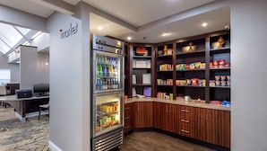 A kitchenette area with a refrigerator stocked with beverages, a microwave, and a coffee machine. There are wooden cabinets above the counter with various food items and condiments. A computer desk with a chair is visible in the background. - Residence Inn by Marriott Little Rock (Little Rock)